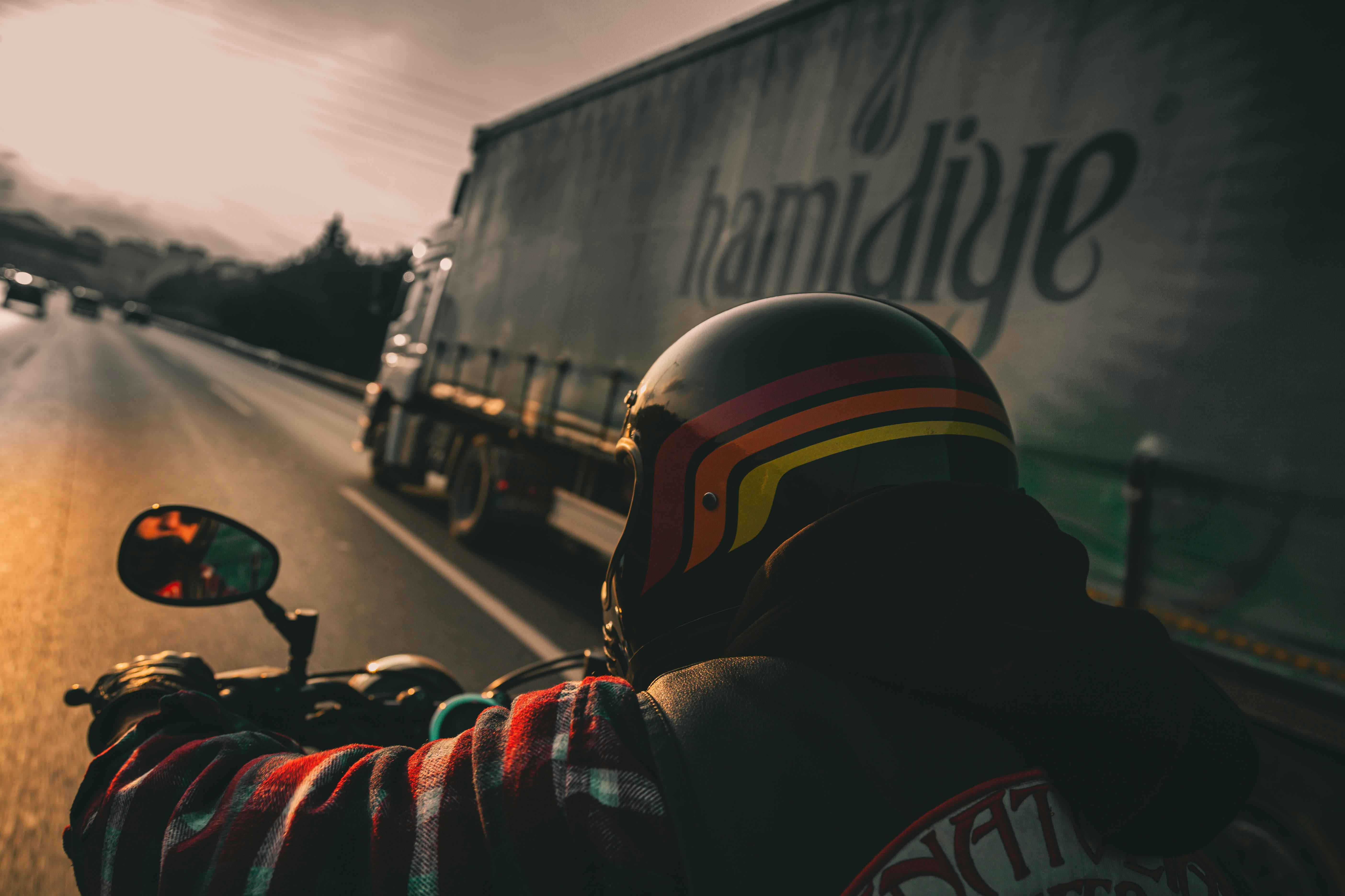 Motorcyclist wearing helmet rides on highway beside a truck at sunset.